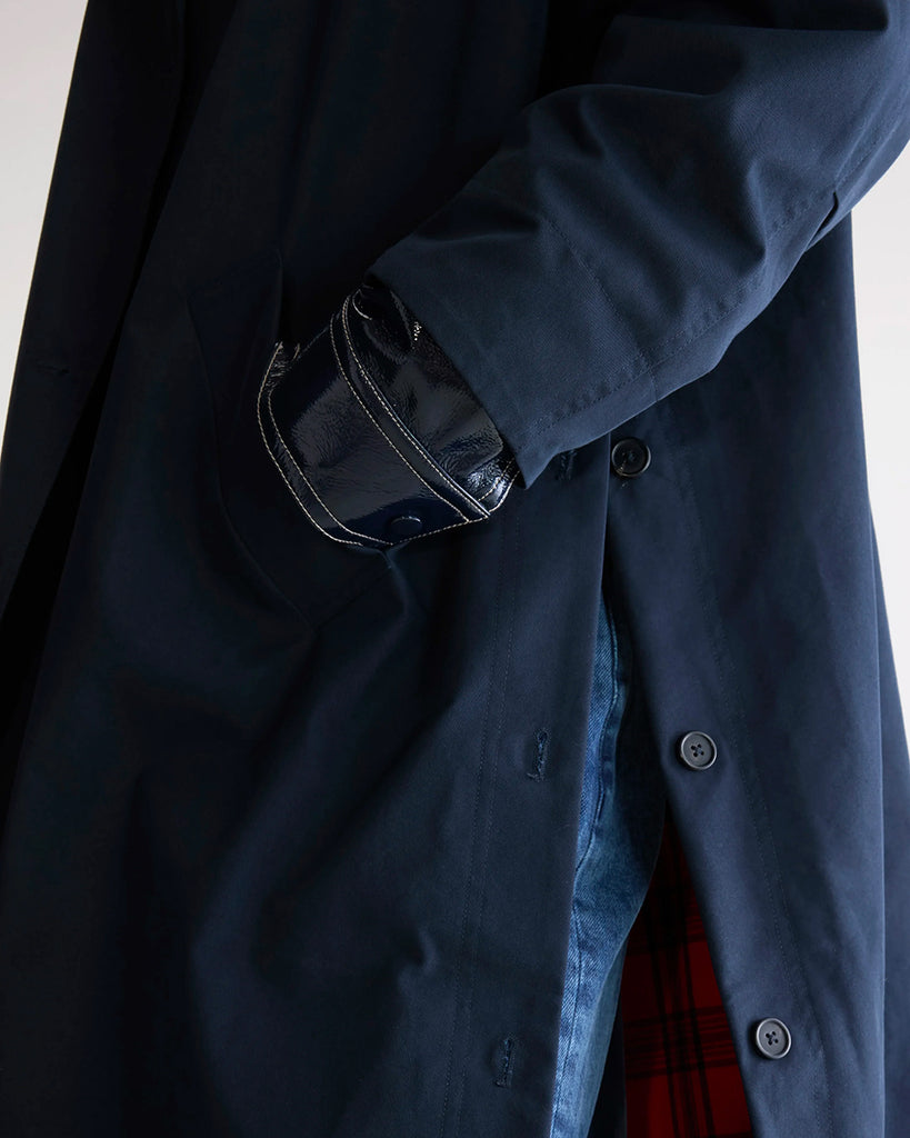 Długi, oversizowy, przejściowy trencz Juttac Bellerose z wysokim kołnierzem, opadającymi ramionami i luźnymi rękawami. Zapinany na jeden, boczny rząd guzików. Płaszcz ma prosty krój i głębokie, boczne rozcięcia zapinane na guziki. Posiada czerwoną podszewkę w kratę. 