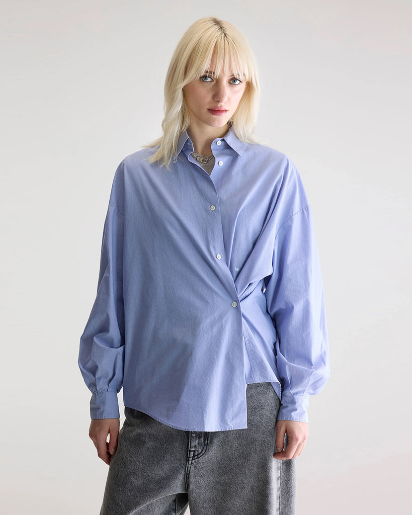 Niebieska, oversizowa koszula w cienkie prążki Gillian Bellerose z opadającymi ramionami i szerokimi rękawami zakończonymi wąskimi mankietami. Zapinana na guziki. Posiada bardzo ciekawą opcję asymetrycznego zapięcia i zwężenia w talii.