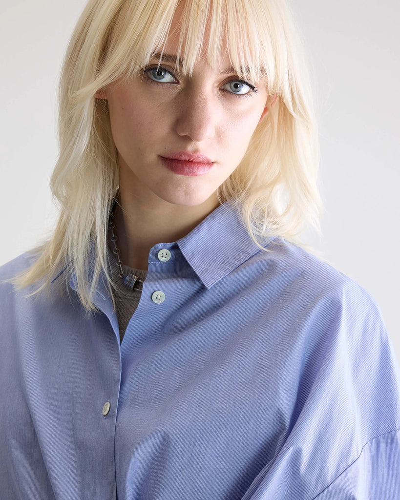 Niebieska, oversizowa koszula w cienkie prążki Gillian Bellerose z opadającymi ramionami i szerokimi rękawami zakończonymi wąskimi mankietami. Zapinana na guziki. Posiada bardzo ciekawą opcję asymetrycznego zapięcia i zwężenia w talii.