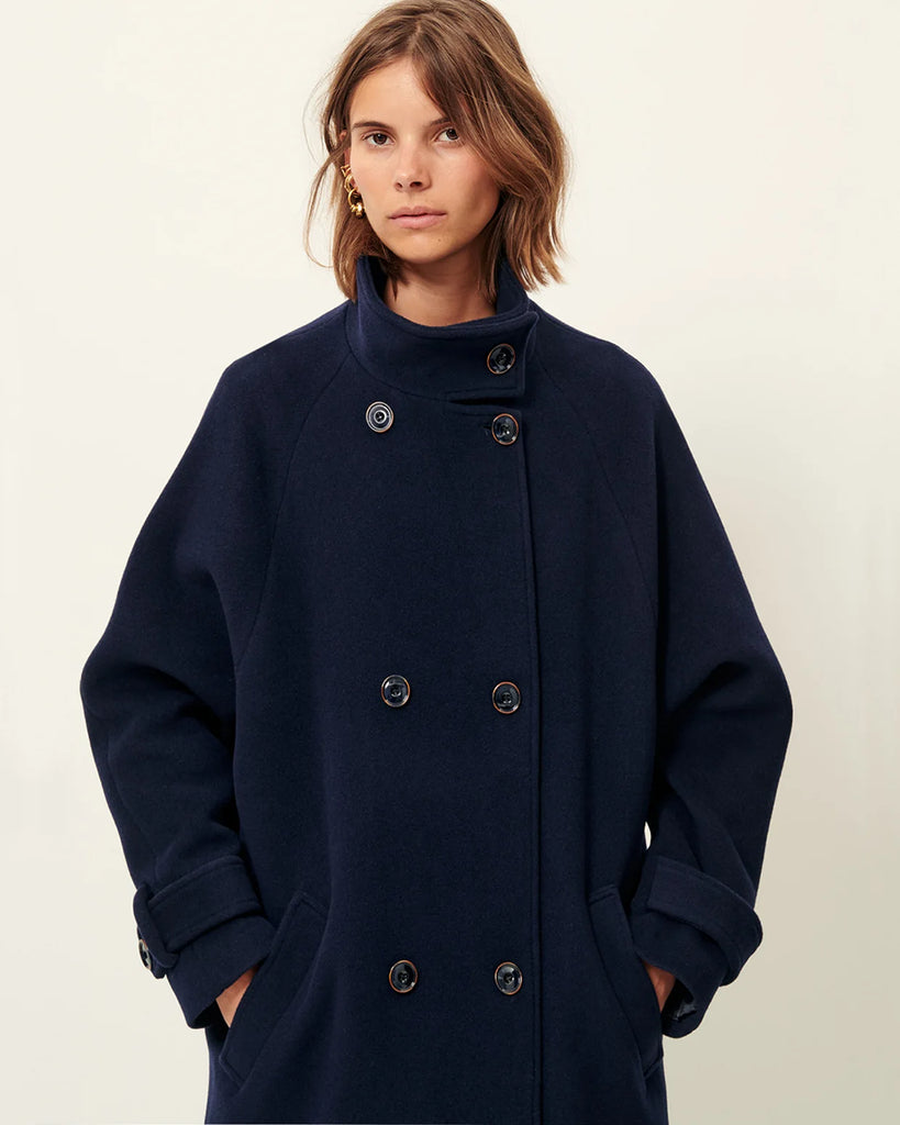 Klasyczny, oversizowy, granatowy płaszcz Bleeckett Indigo o prostym i eleganckim kroju. Ma dwurzędowe zapięcie, z dwoma symetrycznymi rzędami dużych, ciemnych guzików oraz kołnierz typu stójka. Kieszenie wpuszczane po bokach, na wysokości bioder. Sięga mniej więcej do połowy łydki, co nadaje mu klasyczny, zimowy charakter.&nbsp;
