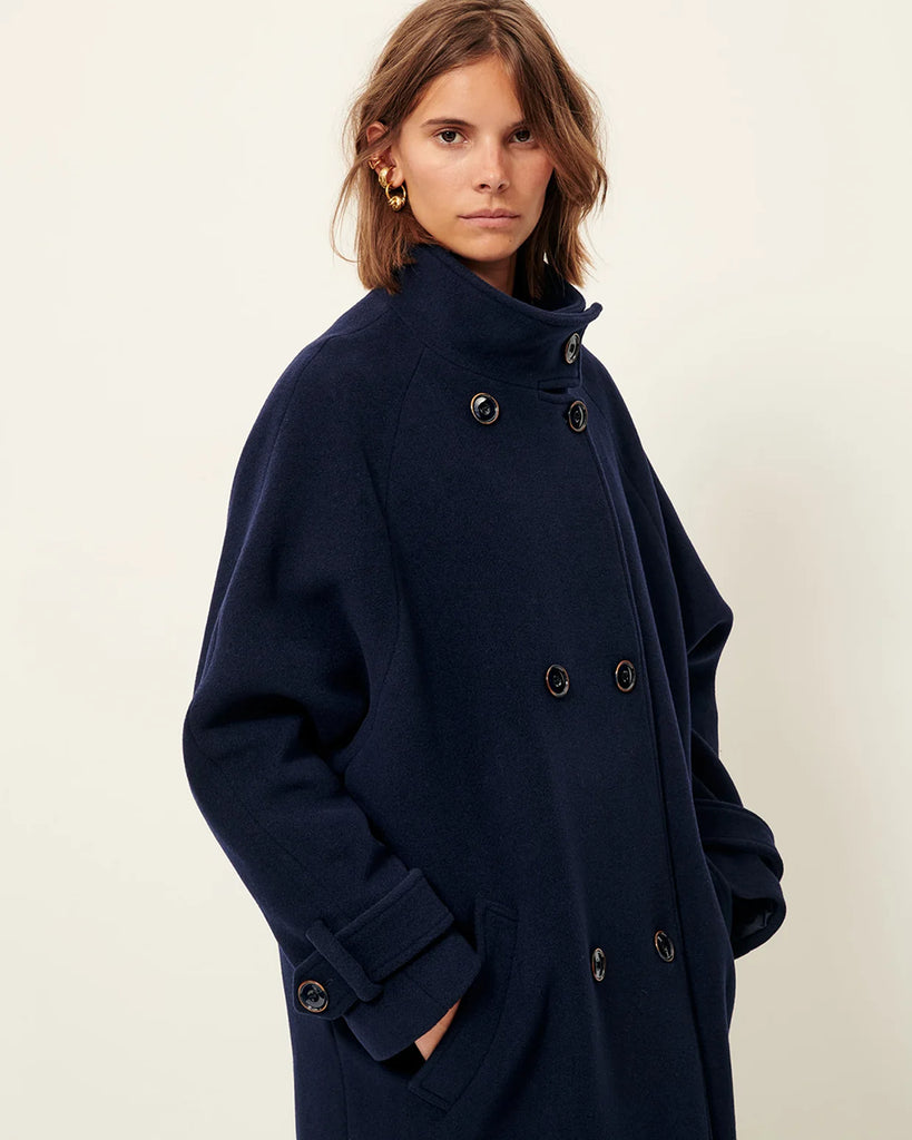 Klasyczny, oversizowy, granatowy płaszcz Bleeckett Indigo o prostym i eleganckim kroju. Ma dwurzędowe zapięcie, z dwoma symetrycznymi rzędami dużych, ciemnych guzików oraz kołnierz typu stójka. Kieszenie wpuszczane po bokach, na wysokości bioder. Sięga mniej więcej do połowy łydki, co nadaje mu klasyczny, zimowy charakter.&nbsp;
