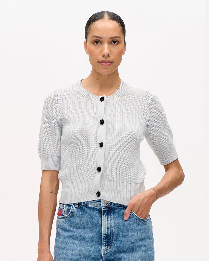 Dzianinowy, krótki kardigan Cathia Grey Melange z krótkim rękawem, o lekko dopasowanym kroju. Zapinany na zatrzaski zdobione błyszczącymi kwiatami. Wykonany z bawełny organicznej. Sweterek zestaw ze swoimi ulubionymi jeansami lub spódnicą.