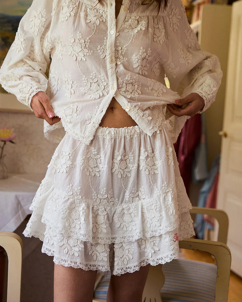 Falbaniaste, kobiece szorty Delphine Off White marki SISSEL EDELBO z elastycznym pasem. Wzdłuż bioder i nogawek ozdobione są falbanami zakończonymi delikatnymi, koronkowymi tasiemkami. Z przodu posiadają haftowany napis 'SISSEL'. Wykonane z bawełny schiffli, zgodnie z ideą marki wykorzystując różnego rodzaju resztki tkanin zamiast produkcji nowych.&nbsp;

-ONE SIZE