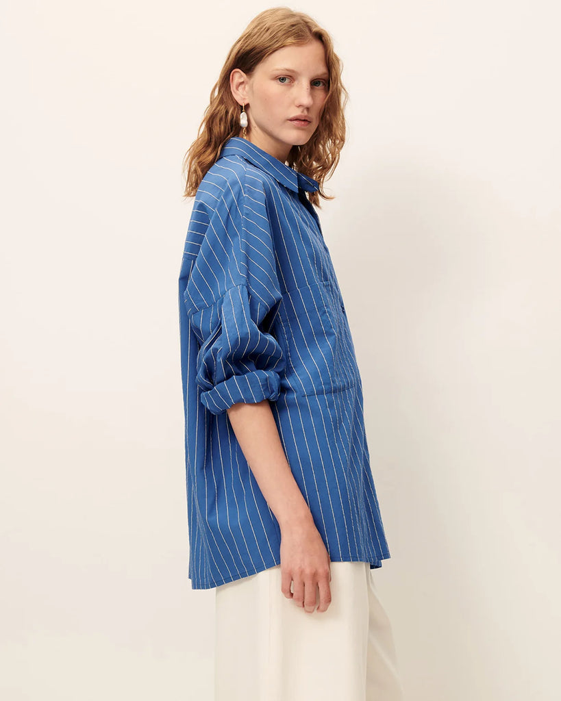 Niebieska luźna koszula Fontania Office Blue z długimi rękawami i opadającymi ramionami. Zapinana na guziki. Z przodu posiada naszywane kieszenie. Stębnowany karczek z tyłu z kontrafałdą i patką na guzik. Wykonana z kompaktowej popeliny w delikatne, teksturowane, dwukolorowe paski.&nbsp;