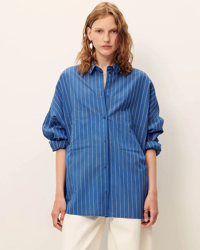 Niebieska luźna koszula Fontania Office Blue z długimi rękawami i opadającymi ramionami. Zapinana na guziki. Z przodu posiada naszywane kieszenie. Stębnowany karczek z tyłu z kontrafałdą i patką na guzik. Wykonana z kompaktowej popeliny w delikatne, teksturowane, dwukolorowe paski.&nbsp;