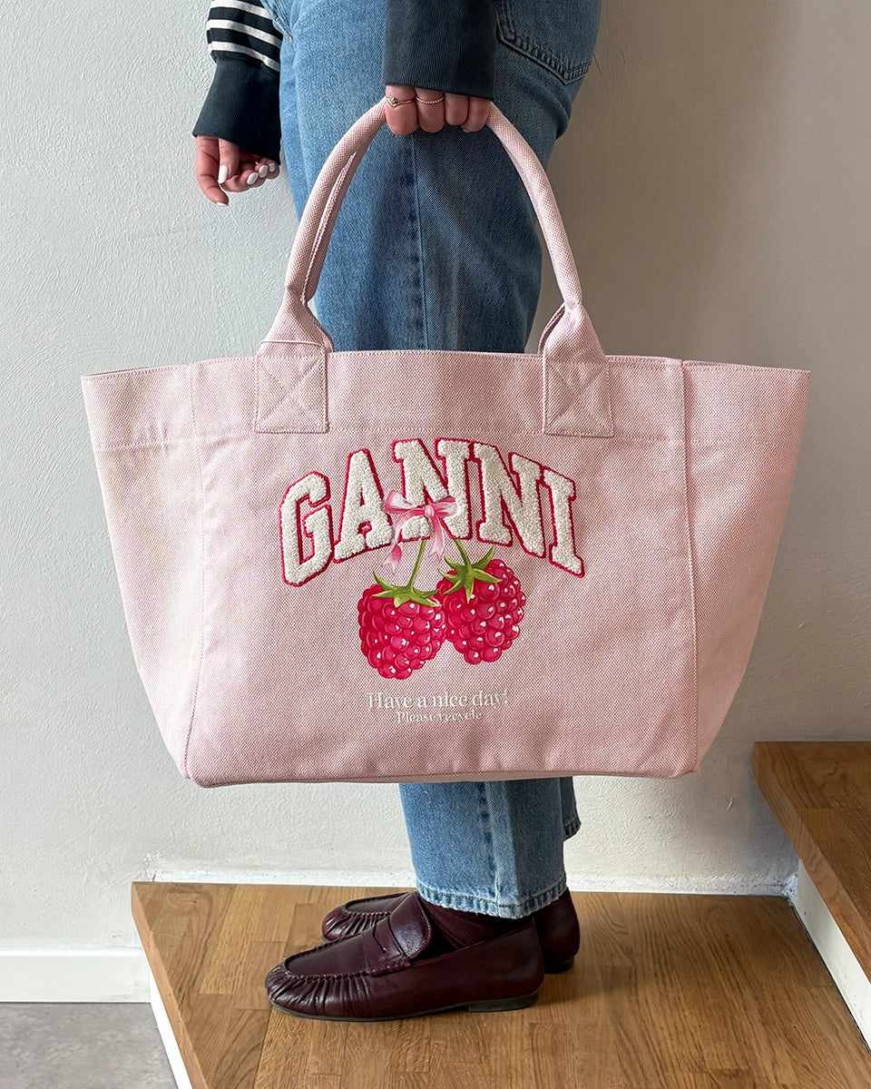 Średniej wielkości, trapezowa, różowa torba shopper 2110 Rasperry GANNI z pluszowym logo marki oraz z uroczym printem z motywem malin. Torba posiada dwie, grube, krótkie rączki do wygodnego trzymania w dłoni oraz odpinany, cienki, długi pasek do wygodnego zawiedzenia na ramieniu. Wewnątrz znajduje się mała kieszonka zapinana na zamek błyskawiczny. Wykonana z grubego, sztywnego, płóciennego materiału.&nbsp;