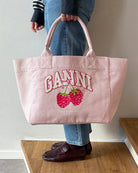 Średniej wielkości, trapezowa, różowa torba shopper 2110 Rasperry GANNI z pluszowym logo marki oraz z uroczym printem z motywem malin. Torba posiada dwie, grube, krótkie rączki do wygodnego trzymania w dłoni oraz odpinany, cienki, długi pasek do wygodnego zawiedzenia na ramieniu. Wewnątrz znajduje się mała kieszonka zapinana na zamek błyskawiczny. Wykonana z grubego, sztywnego, płóciennego materiału.&nbsp;