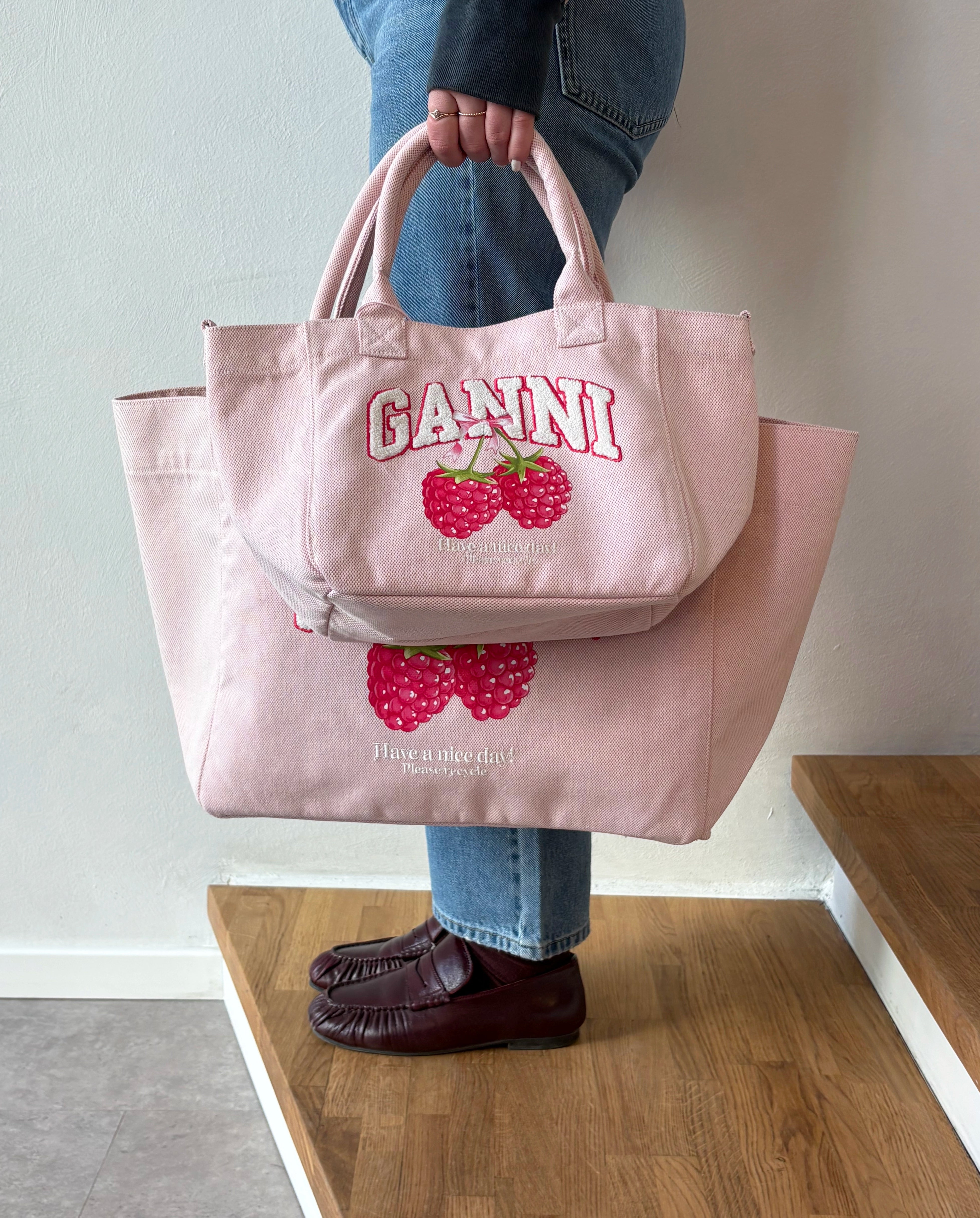 Średniej wielkości, trapezowa, różowa torba shopper 2110 Rasperry GANNI z pluszowym logo marki oraz z uroczym printem z motywem malin. Torba posiada dwie, grube, krótkie rączki do wygodnego trzymania w dłoni oraz odpinany, cienki, długi pasek do wygodnego zawiedzenia na ramieniu. Wewnątrz znajduje się mała kieszonka zapinana na zamek błyskawiczny. Wykonana z grubego, sztywnego, płóciennego materiału.&nbsp;