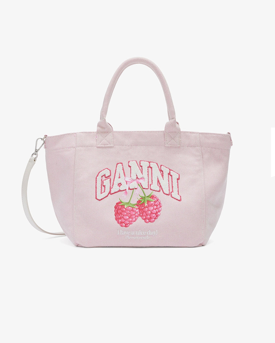 Średniej wielkości, trapezowa, różowa torba shopper 2110 Rasperry GANNI z pluszowym logo marki oraz z uroczym printem z motywem malin. Torba posiada dwie, grube, krótkie rączki do wygodnego trzymania w dłoni oraz odpinany, cienki, długi pasek do wygodnego zawiedzenia na ramieniu. Wewnątrz znajduje się mała kieszonka zapinana na zamek błyskawiczny. Wykonana z grubego, sztywnego, płóciennego materiału.&nbsp;