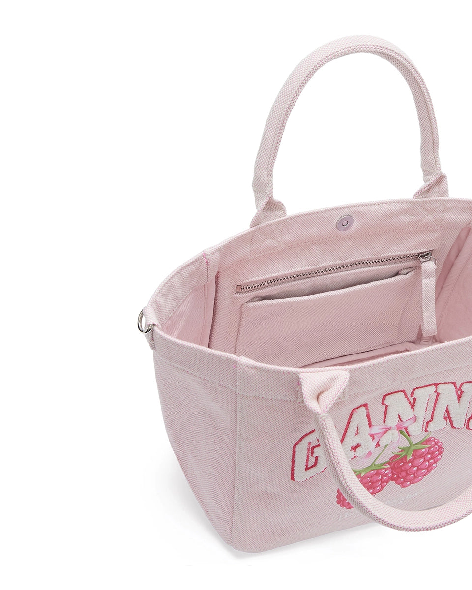 Średniej wielkości, trapezowa, różowa torba shopper 2110 Rasperry GANNI z pluszowym logo marki oraz z uroczym printem z motywem malin. Torba posiada dwie, grube, krótkie rączki do wygodnego trzymania w dłoni oraz odpinany, cienki, długi pasek do wygodnego zawiedzenia na ramieniu. Wewnątrz znajduje się mała kieszonka zapinana na zamek błyskawiczny. Wykonana z grubego, sztywnego, płóciennego materiału.&nbsp;