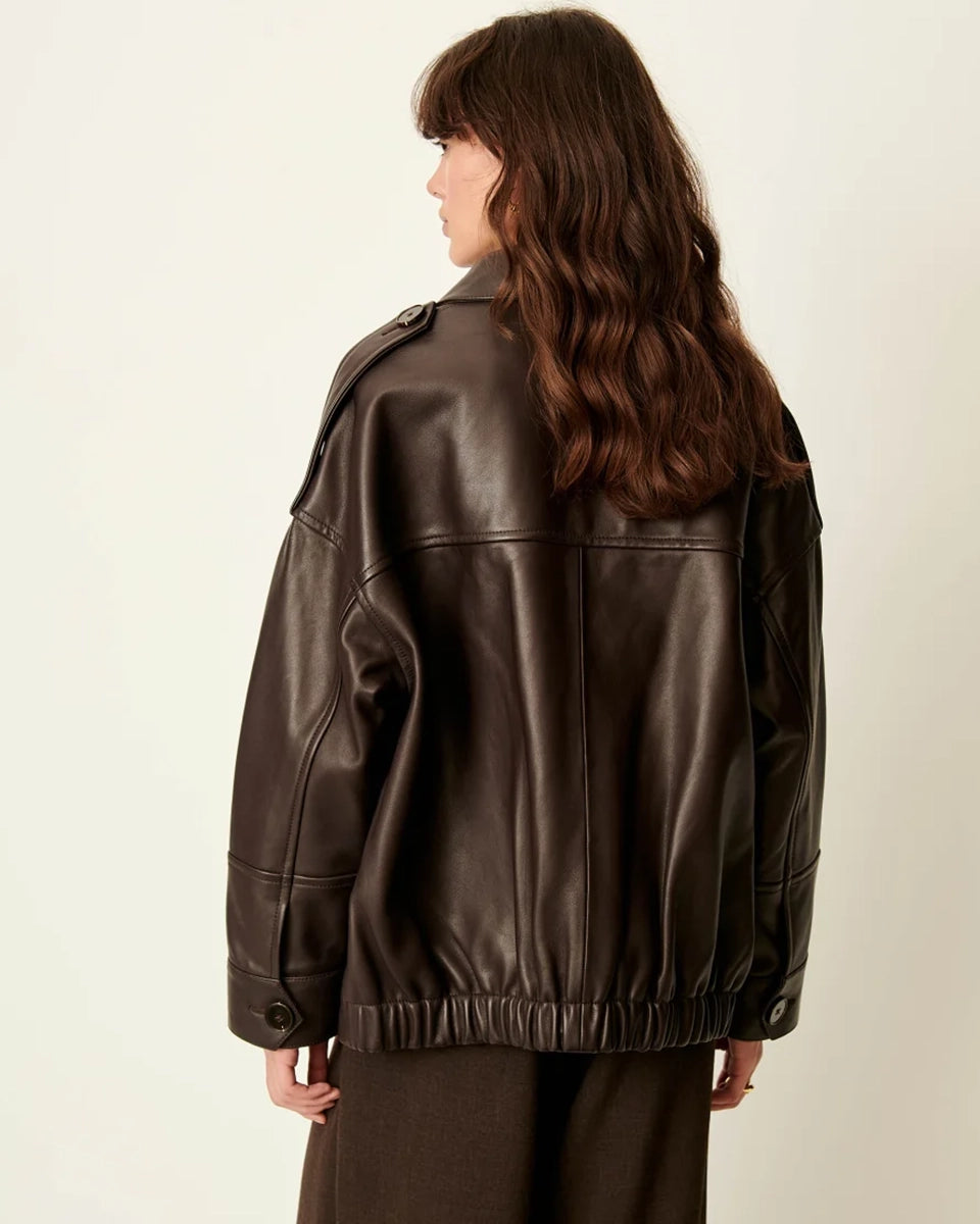 Brązowa, luźna kurtka Harriboyd Coffee Leather francuskiej marki Sessun w stylu klasycznej pilotki. Wykonana jest z gładkiej, ciemnobrązowej skóry o lekkim połysku. Z przodu zapinana jest na duże, czarne guziki, a po bokach ma duże naszywane kieszenie. Rękawy są długie, z mankietami zapinanymi na guzik