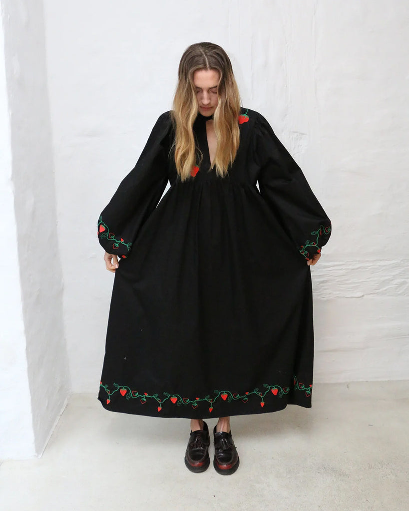 Oversizowa sukienka maxi Sigrid HELMSTEDT w uroczy haft z motywem truskawek. Sukienka ma rozszerzane, długie rękawy i okrągły dekolt z trójkątnym wycięciem i wiązaniem. Jest odcięta pod biustem. Sukienka ma długość do kostek. 