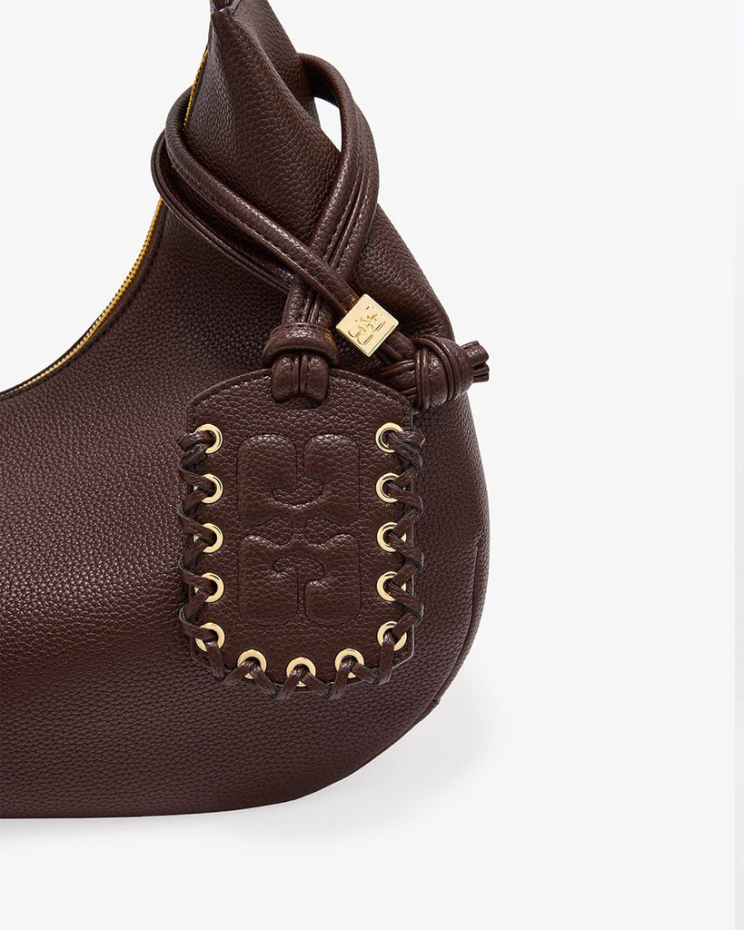 Duża, brązowa torba o kształcie półksiężyca 7298 Hobo Medium GANNI z dużą, szeroką rączką do wygodnego zawieszenia na ramieniu. Z przodu posiada niewielki wypukły logotyp marki. Z tyłu, u dołu znajduje się wytłaczany napis GANNI. Do torby dołączona jest duża, lustrzana zawieszka. Jest zapinana za pomocą zamka błyskawicznego. Wewnątrz posiada niewielką kieszonkę na najważniejsze drobiazgi takie jak klucze czy karta kredytowa. Wykonana z opatentowanego przez markę, ekologicznego materiału bedącego połączeniem