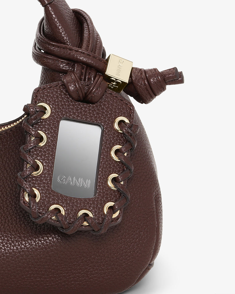 Mała, brązowa torebka w kształcie półksiężyca Hobo Mini Chicory Coffee z uchwytem i regulowanym, odpinanym paskiem oraz zapięciem na suwak. Z przodu posiada niewielki wypukły logotyp marki. Z tyłu, u dołu znajduje się wytłaczany napis GANNI. Do torebki dołączona jest lustrzana zawieszka. Wewnątrz posiada niewielką kieszonkę na najważniejsze drobiazgi takie jak klucze czy karta kredytowa.