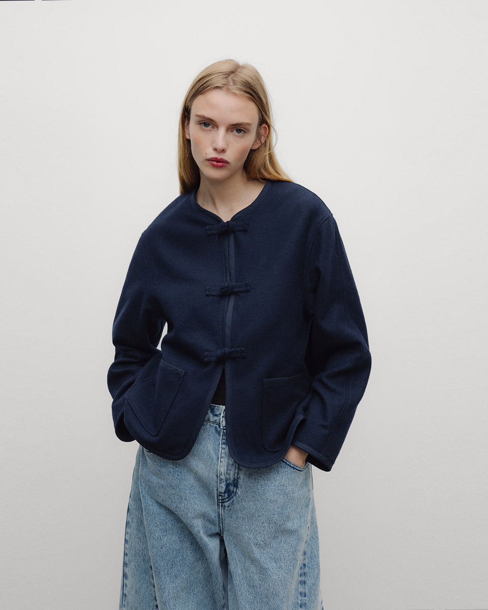Elegancka, ciemnogranatowa, krótka kurtka Indie Black Iris o fasonie inspirowanym stylem wschodnim. Model ma prosty krój z okrągłym dekoltem bez kołnierzyka oraz długie rękawy o swobodnej linii. Charakterystycznym elementem są ozdobne, tekstylne zapięcia typu chińskiego, zwane węzełkami, które biegną wzdłuż przedniej plisy. Kurtka posiada również dwie duże, naszywane kieszenie oraz zaokrąglone doły po bokach, co nadaje jej miękki, nowoczesny wygląd. Wykonana z mieszanki z wełną.

-Inspirowana stylem wschodn