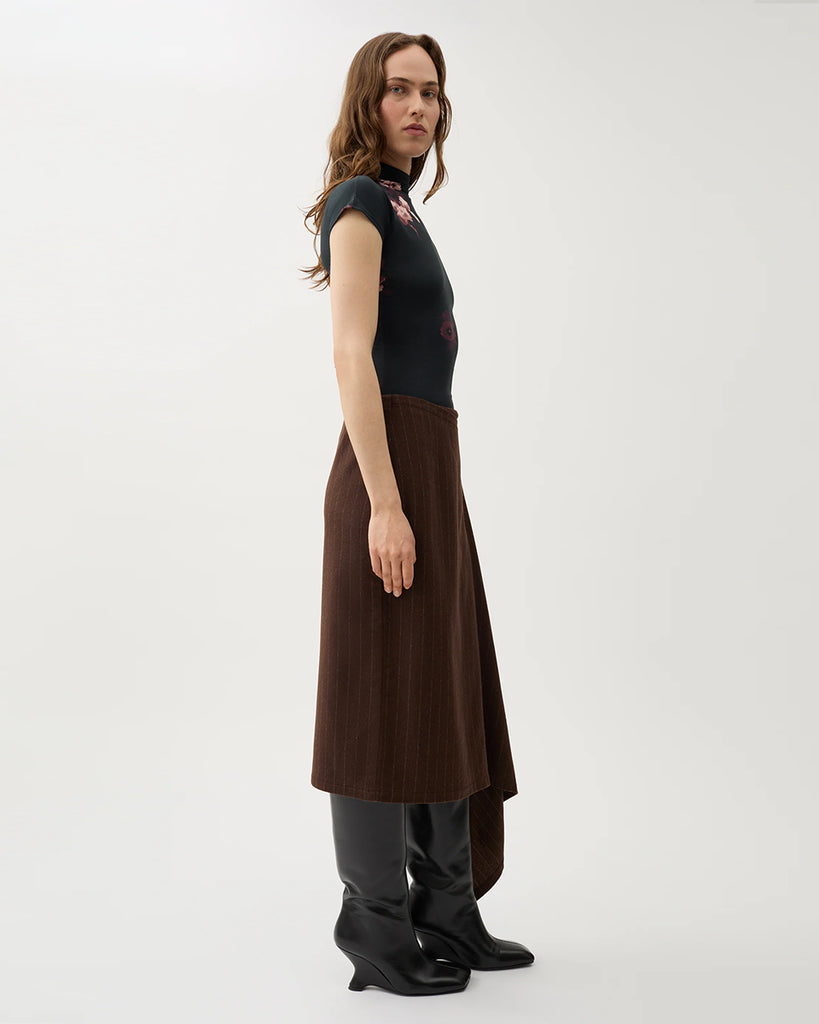 Kopertowa, elegancka spódnica Josefina Chicory Coffee o asymetrycznym kroju w kolorze brązowym w paski. Po lewej stronie ma dłuższy, opadający fragment materiału, tworzący efekt skośnego, nieregularnego dołu. Na górze, z przodu znajduje się ozdobny guzik. Długość sięga za kolano.&nbsp;