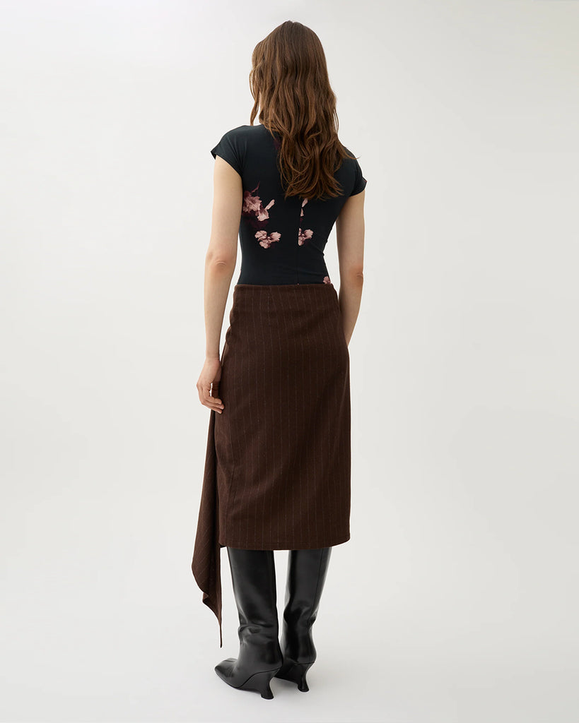 Kopertowa, elegancka spódnica Josefina Chicory Coffee o asymetrycznym kroju w kolorze brązowym w paski. Po lewej stronie ma dłuższy, opadający fragment materiału, tworzący efekt skośnego, nieregularnego dołu. Na górze, z przodu znajduje się ozdobny guzik. Długość sięga za kolano.&nbsp;