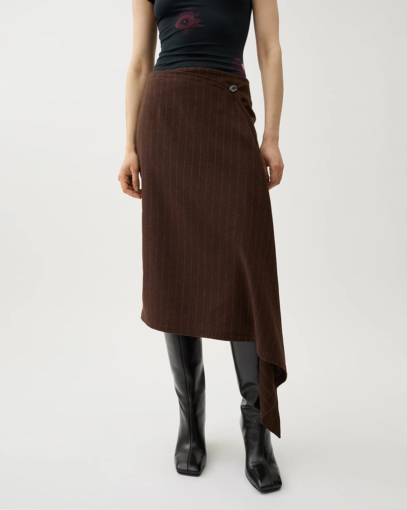 Kopertowa, elegancka spódnica Josefina Chicory Coffee o asymetrycznym kroju w kolorze brązowym w paski. Po lewej stronie ma dłuższy, opadający fragment materiału, tworzący efekt skośnego, nieregularnego dołu. Na górze, z przodu znajduje się ozdobny guzik. Długość sięga za kolano.&nbsp;