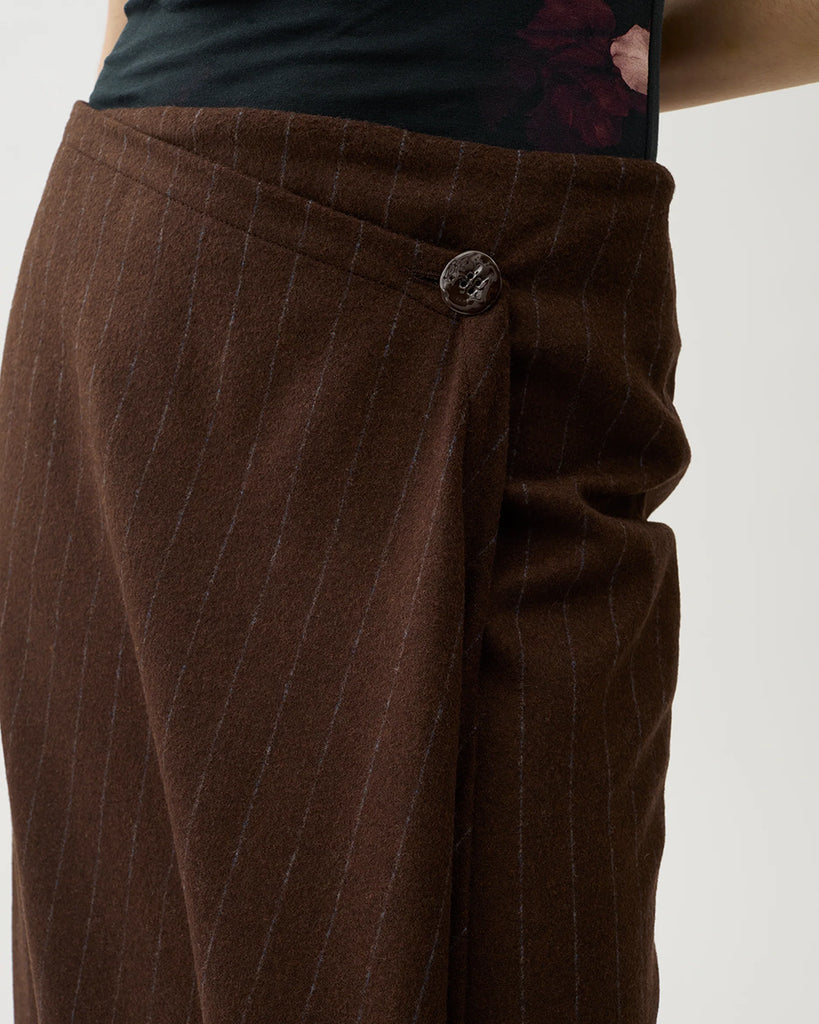 Kopertowa, elegancka spódnica Josefina Chicory Coffee o asymetrycznym kroju w kolorze brązowym w paski. Po lewej stronie ma dłuższy, opadający fragment materiału, tworzący efekt skośnego, nieregularnego dołu. Na górze, z przodu znajduje się ozdobny guzik. Długość sięga za kolano.&nbsp;