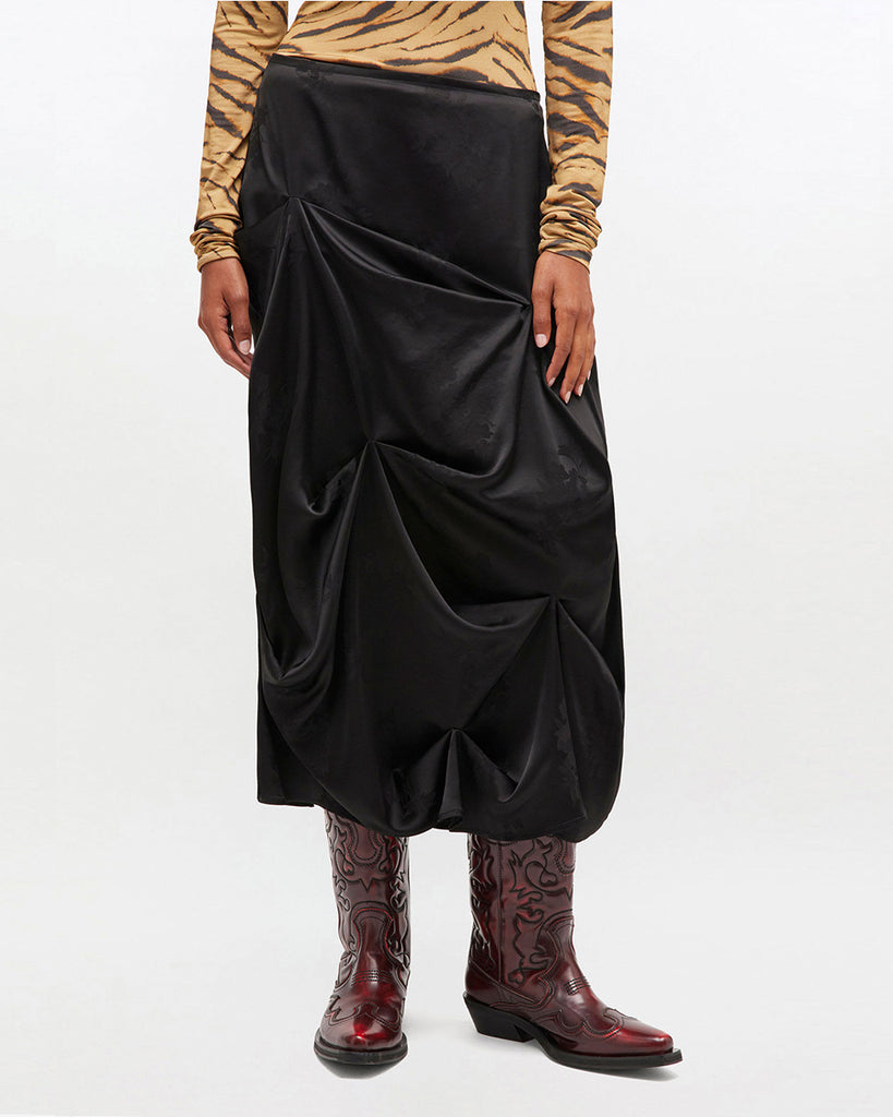 Luźna, drapowana spódnica 0997 Ganni Satin Black o satynowym wyglądzie, zdobiona kwiatowymi wzorami. Ma długość do połowy łydki. Zapinana z boku na zamek błyskawiczny. Idealna do luźnego topu i butów na obcasie typu kowbojki.&nbsp;