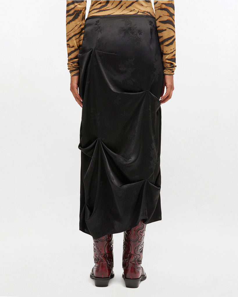 Luźna, drapowana spódnica 0997 Ganni Satin Black o satynowym wyglądzie, zdobiona kwiatowymi wzorami. Ma długość do połowy łydki. Zapinana z boku na zamek błyskawiczny. Idealna do luźnego topu i butów na obcasie typu kowbojki.&nbsp;