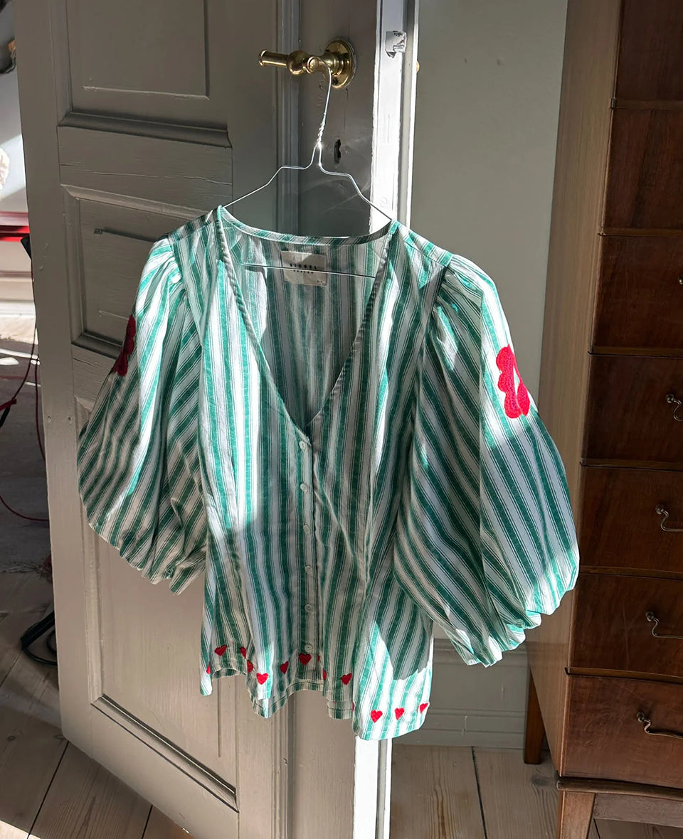 Kobieca bluzka koszulowa Lotta Green Stripes SISSEL EDELBO z dopasowaną talią i marszczeniem z tyłu. Ma głęboki dekolt w serek i zapięcie z przodu na guziki z masy perłowej. Rękawy mają długość 3/4 i zakończone są gumką. Ozdobiona jest delikatnym haftem kwiatowym i serduszkami. Wykonana z organicznej bawełny popelinowej w paski.&nbsp;

-Dopasowana talia

-100% bawełna organiczna

-Zamów bluzkę w swoim standardowym rozmiarze&nbsp;