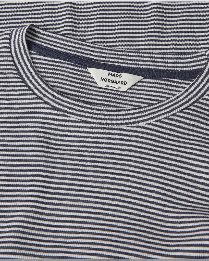 Minimalistyczny longsleeve 2x2 Tuba MADS NORGAARD w cienkie, granatowo-kremowe paski. Bluzka ma okrągły dekolt, przylegający fason i wąskie, długie rękawy. Wykonany jest z elastycznej, bawełnianej tkaniny z prążkowaną fakturą