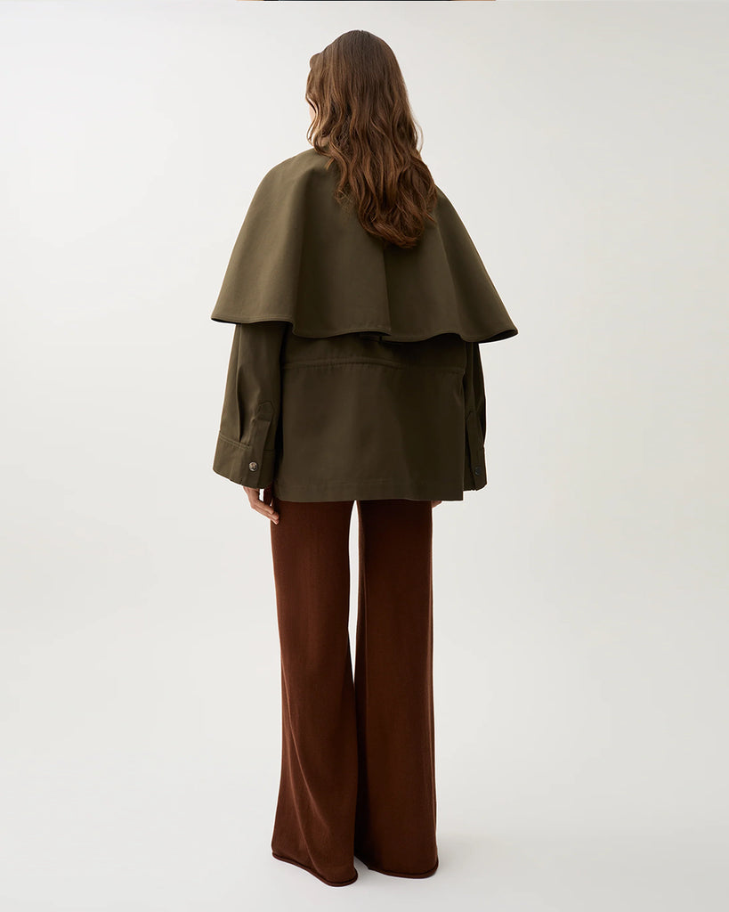 Minimalistyczna, pudełkowa kurtka Malin Brown Moss typu pelerynowa w ciepłym, oliwkowo-brązowym kolorze. Górna część (peleryna) jest odpinana, co pozwala nosić kurtkę na dwa sposoby. Ma prosty, koszulowy kołnierz, zapięcia na napy i duże naszywane kieszenie z przodu.