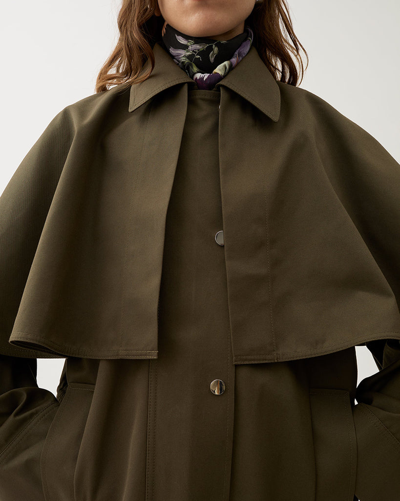 Minimalistyczna, pudełkowa kurtka Malin Brown Moss typu pelerynowa w ciepłym, oliwkowo-brązowym kolorze. Górna część (peleryna) jest odpinana, co pozwala nosić kurtkę na dwa sposoby. Ma prosty, koszulowy kołnierz, zapięcia na napy i duże naszywane kieszenie z przodu.