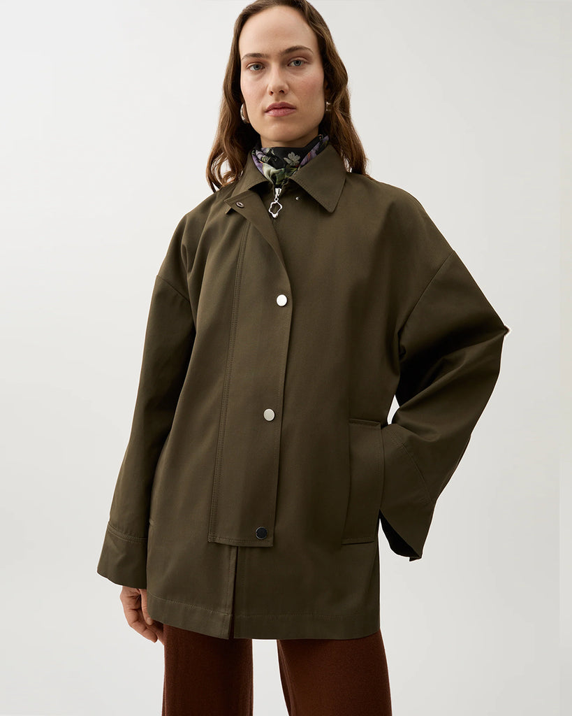 Minimalistyczna, pudełkowa kurtka Malin Brown Moss typu pelerynowa w ciepłym, oliwkowo-brązowym kolorze. Górna część (peleryna) jest odpinana, co pozwala nosić kurtkę na dwa sposoby. Ma prosty, koszulowy kołnierz, zapięcia na napy i duże naszywane kieszenie z przodu.