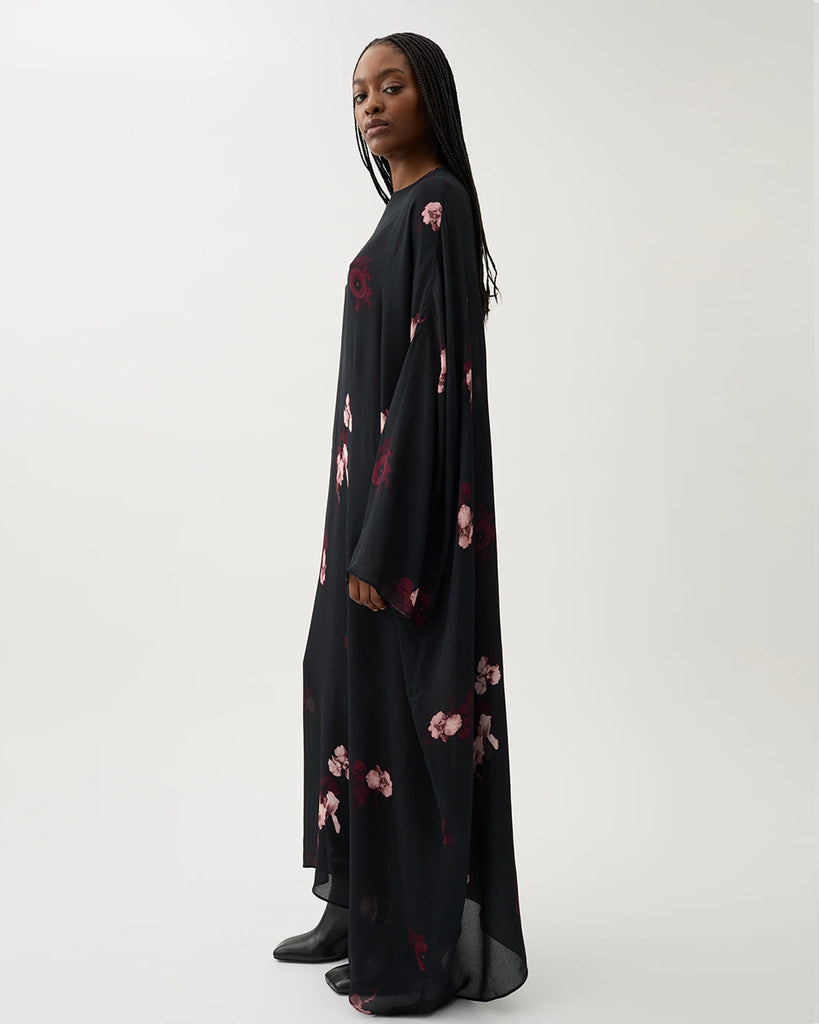 Długa, luźna sukienka Marge Wine Black w kolorze czarnym z kwiatowym printem. Ma okrągły dekolt z wiązaniem z tyłu oraz raglanowe, przedłużone rękawy. Dół sukienki jest asymetryczny.&nbsp;