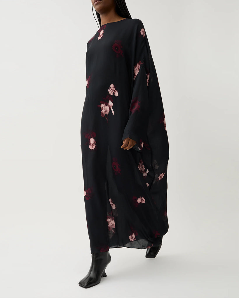 Długa, luźna sukienka Marge Wine Black w kolorze czarnym z kwiatowym printem. Ma okrągły dekolt z wiązaniem z tyłu oraz raglanowe, przedłużone rękawy. Dół sukienki jest asymetryczny.&nbsp;