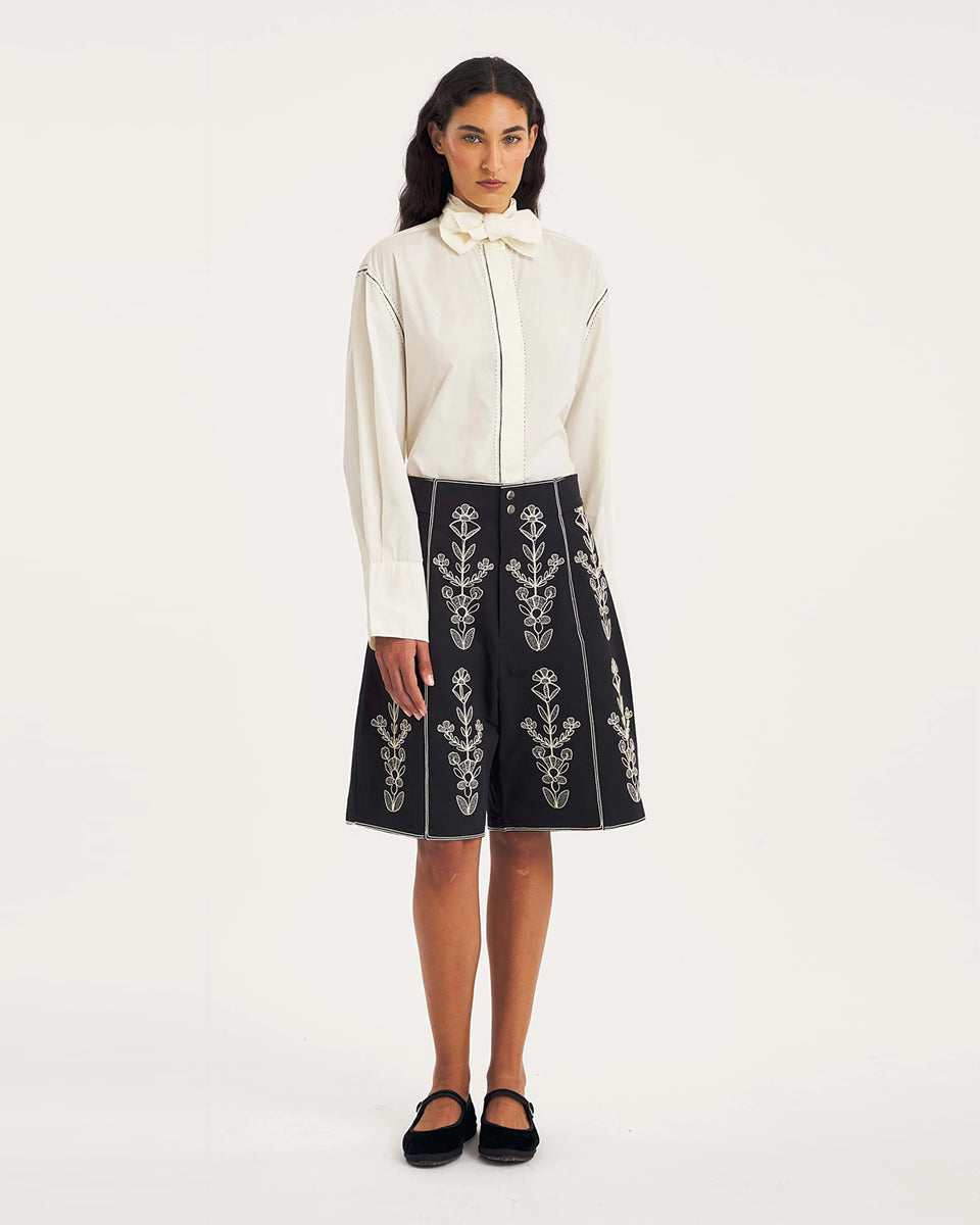 Dłuższe, eleganckie szorty typu bermudy Maria Black marki Antik Batik sięgające do kolan. Mają luźne, szerokie i prosto ścięte nogawki oraz wysoki stan z szerokim pasem i szlufkami. Zapinane są z przodu na dwa jasne guziki umieszczone pionowo. Wzdłuż nogawek oraz wokół kieszeni i paska mają kontrastowe, jasne lamówki. Przód, jak i tył zdobią bogate białe hafty o motywie roślinnym. Hafty umieszczone są symetrycznie w pionowych rzędach, co nawiązuje do tradycyjnego stylu boho.&nbsp;

-Luźne, proste nogawki

-
