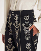 Dłuższe, eleganckie szorty typu bermudy Maria Black marki Antik Batik sięgające do kolan. Mają luźne, szerokie i prosto ścięte nogawki oraz wysoki stan z szerokim pasem i szlufkami. Zapinane są z przodu na dwa jasne guziki umieszczone pionowo. Wzdłuż nogawek oraz wokół kieszeni i paska mają kontrastowe, jasne lamówki. Przód, jak i tył zdobią bogate białe hafty o motywie roślinnym. Hafty umieszczone są symetrycznie w pionowych rzędach, co nawiązuje do tradycyjnego stylu boho.&nbsp;

-Luźne, proste nogawki

-
