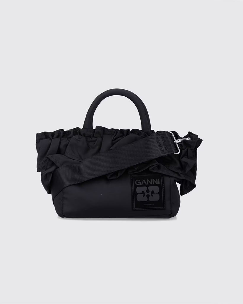 Torba typu tote 7187 Mini Shopper Black marki GANNI z uchwytami i paskiem na ramię. Posiada jedną dużą komorę z wewnętrzną kieszenią zapinaną na zamek. U dołu znajduje się niewielkie logo marki. Wyjątkowości dodaje zdobienie w postaci falbanek. Zaprojektowana, aby pomieścić najpotrzebniejsze drobiazgi.