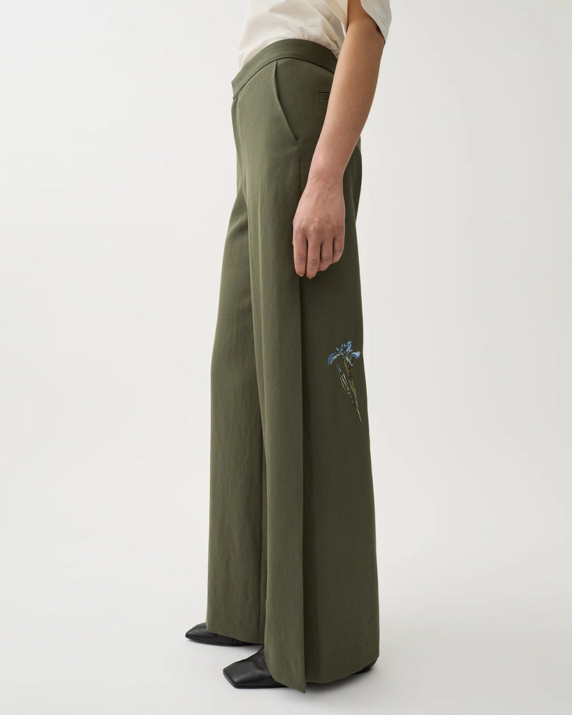 Luźne, garniturowe spodnie Nora Dark Moss w kolorze oliwkowej zieleni z haftowanymi zdobieniami w postaci kwiatów, wysokim stanem i kantem. Posiadają długą, rozszerzaną nogawkę. Mają elastyczny pas w talii i boczne kieszenie. Wykonane z lejącego, dzianinowego materiału zapewniającego komfort i swobodę ruchów. Zestaw je z marynarką Stand Dark Moss tej samej marki.
