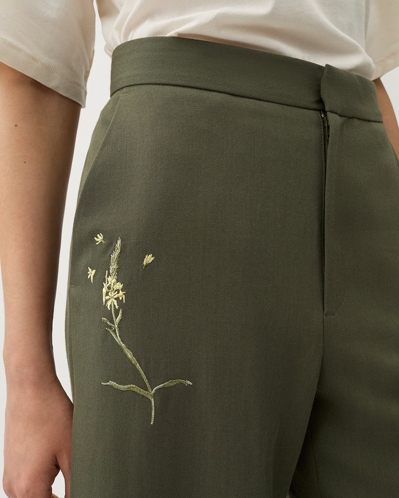 Luźne, garniturowe spodnie Nora Dark Moss w kolorze oliwkowej zieleni z haftowanymi zdobieniami w postaci kwiatów, wysokim stanem i kantem. Posiadają długą, rozszerzaną nogawkę. Mają elastyczny pas w talii i boczne kieszenie. Wykonane z lejącego, dzianinowego materiału zapewniającego komfort i swobodę ruchów. Zestaw je z marynarką Stand Dark Moss tej samej marki.
