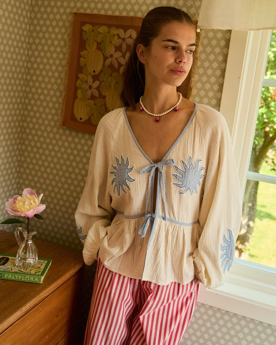 Luźna, kobieca bluzka Ramona Off White marki SISSEL EDELBO wykonana z miękkiej, waflowej tkaniny o podwójnej fakturze, wykonanej z bawełny organicznej. Posiada odcięcie w talii z marszczeniami, które tworzą sylwetkę z falbanką. Ma szerokie rękawy typu raglan z marszczeniami na ramionach i elastycznymi zakończeniami rękawów. Posiada głęboki dekolt w serek i dwa sznurki do wiązania z przodu. Ozdobiony jest haftem z motywem słońca i koralikami z masy perłowej.&nbsp;

-ONE SIZE