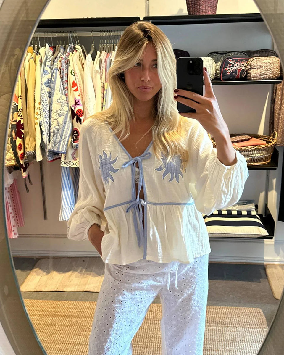 Luźna, kobieca bluzka Ramona Off White marki SISSEL EDELBO wykonana z miękkiej, waflowej tkaniny o podwójnej fakturze, wykonanej z bawełny organicznej. Posiada odcięcie w talii z marszczeniami, które tworzą sylwetkę z falbanką. Ma szerokie rękawy typu raglan z marszczeniami na ramionach i elastycznymi zakończeniami rękawów. Posiada głęboki dekolt w serek i dwa sznurki do wiązania z przodu. Ozdobiony jest haftem z motywem słońca i koralikami z masy perłowej.&nbsp;

-ONE SIZE