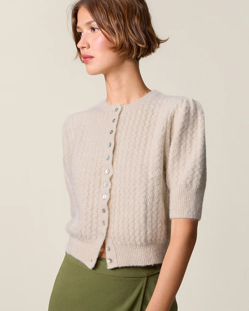 Krótki kardigan z bufiastymi rękawami przed łokieć Cassia Rouje. Posiada okrągły dekolt. Zapinany na drobne guziki z masy perłowej. Sweter ma prosty krój i jest wykończony ściągaczami.