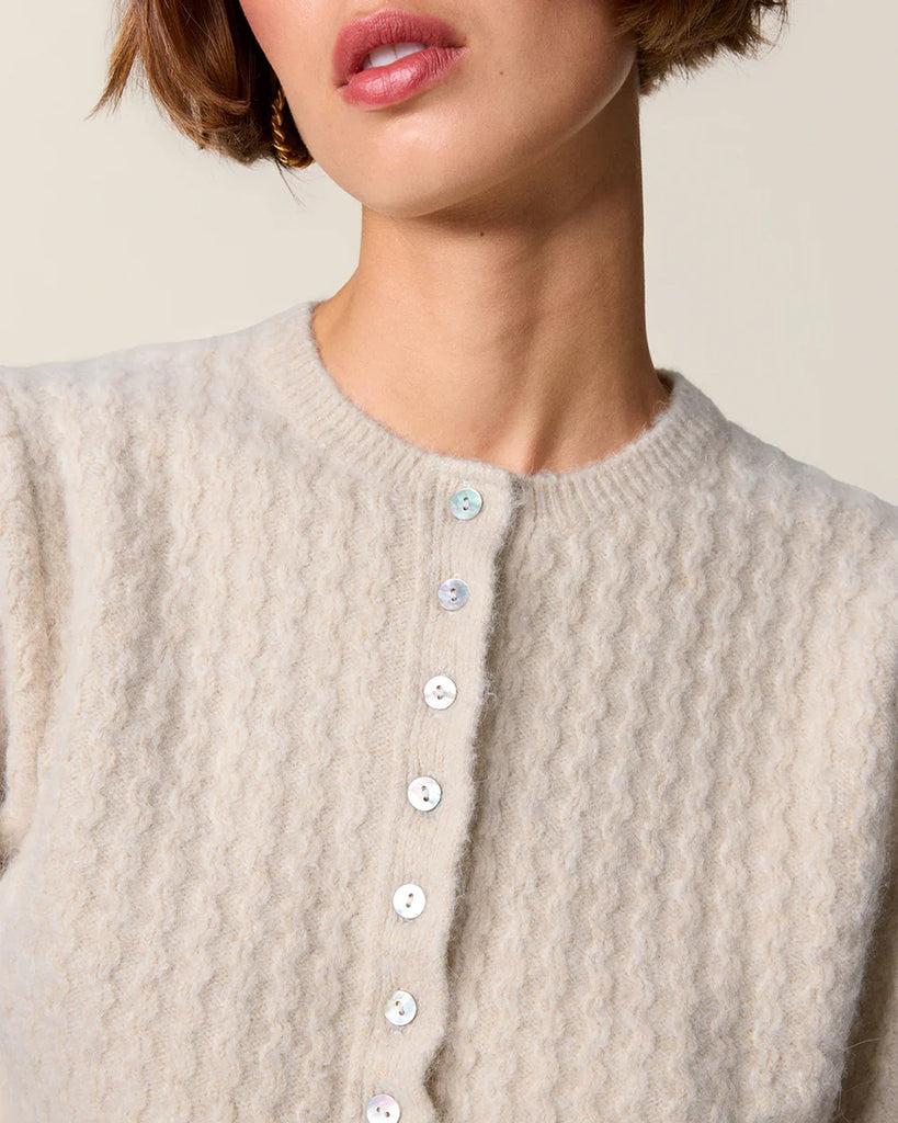 Krótki kardigan z bufiastymi rękawami przed łokieć Cassia Rouje. Posiada okrągły dekolt. Zapinany na drobne guziki z masy perłowej. Sweter ma prosty krój i jest wykończony ściągaczami.