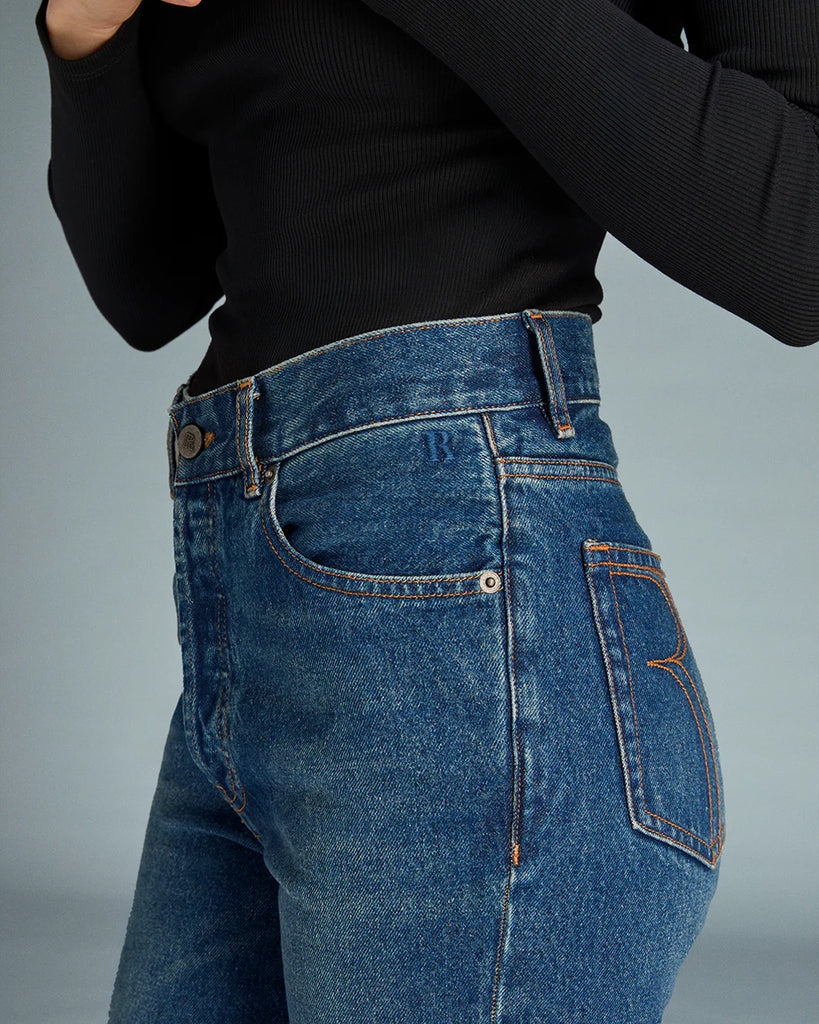 Granatowe jeansy o kobiecym fasonie Concorde Rouje z wysokim stanem i rozszerzaną u dołu, skróconą nogawką. Dopasowane w biodrach i udach. Posiadają pięć kieszeni. Nad lewą kieszenią znajduje się niewielka, haftowana litera R. Zapinane na zamek błyskawiczny i guzik. To model pięknie podkreślający sylwetkę. 