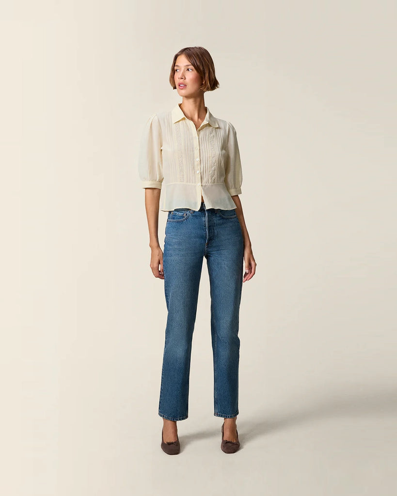 Luźna, kremowa bluzka Dorothea Rouje z krótkim, bufiastym rękawem i kołnierzykiem. Plisowana i haftowana na dekolcie. Zapinana na guziki z masy perłowej. Lekko wcięta w talii. Wykonana z cienkiej i zwiewnej tkaniny z bawełny i wiskozy