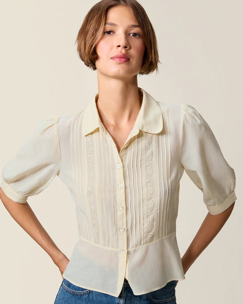 Luźna, kremowa bluzka Dorothea Rouje z krótkim, bufiastym rękawem i kołnierzykiem. Plisowana i haftowana na dekolcie. Zapinana na guziki z masy perłowej. Lekko wcięta w talii. Wykonana z cienkiej i zwiewnej tkaniny z bawełny i wiskozy