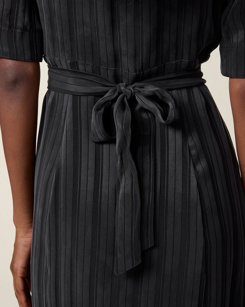 Czarna sukienka midi w paski Laureline Rouje z krótkimi, bufiastymi rękawami, trójkątnym dekoltem i kołnierzykiem. Zapinana na guziki. Dopasowana i wiązana z tyłu w talii za pomocą paska. Sukienka ma długość do połowy łydki.