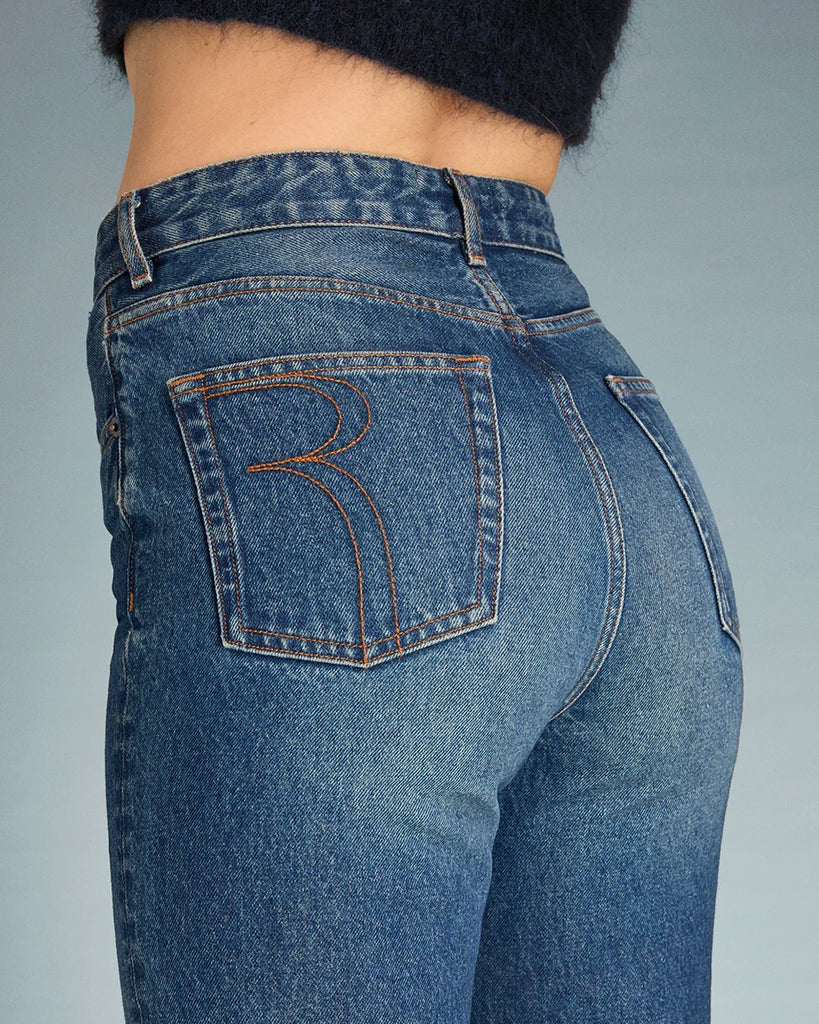 Granatowe jeansy o kobiecym fasonie Madeleine Rouje z wysokim stanem i rozszerzaną u dołu nogawką. Dopasowane w biodrach i udach. Posiadają pięć kieszeni. Nad lewą kieszenią znajduje się niewielka, haftowana litera R. Zapinane na zamek błyskawiczny i guzik.