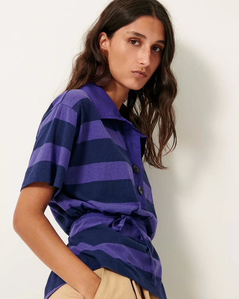 Luźny t-shirt polo Polette Sessun w szerokie, fioletowe paski. Bluzka posiada kołnierzyk i krótki rękaw. Jest wiązana w talii za pomocą cienkiego, wyciąganego sznurka. Pod szyją zapinany na guziki.