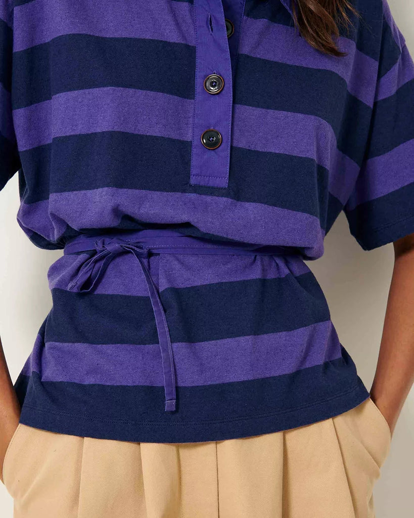 Luźny t-shirt polo Polette Sessun w szerokie, fioletowe paski. Bluzka posiada kołnierzyk i krótki rękaw. Jest wiązana w talii za pomocą cienkiego, wyciąganego sznurka. Pod szyją zapinany na guziki.