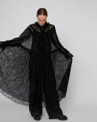 Długa, przezroczysta sukienka Sheer Lace Maxi z koronki z cekinami. Ma zabudowany, efektowny dekolt oraz długie rękawy. Z przodu, na długości ma zapięcia. Można ją nosić zapięta jako sukienkę, bądź rozpiętą jako pelerynę. Sukienka nie posiada podszewki.&nbsp;