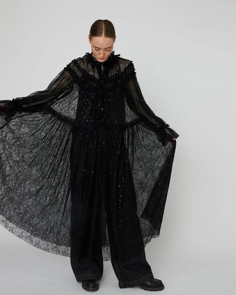 Długa, przezroczysta sukienka Sheer Lace Maxi z koronki z cekinami. Ma zabudowany, efektowny dekolt oraz długie rękawy. Z przodu, na długości ma zapięcia. Można ją nosić zapięta jako sukienkę, bądź rozpiętą jako pelerynę. Sukienka nie posiada podszewki.&nbsp;